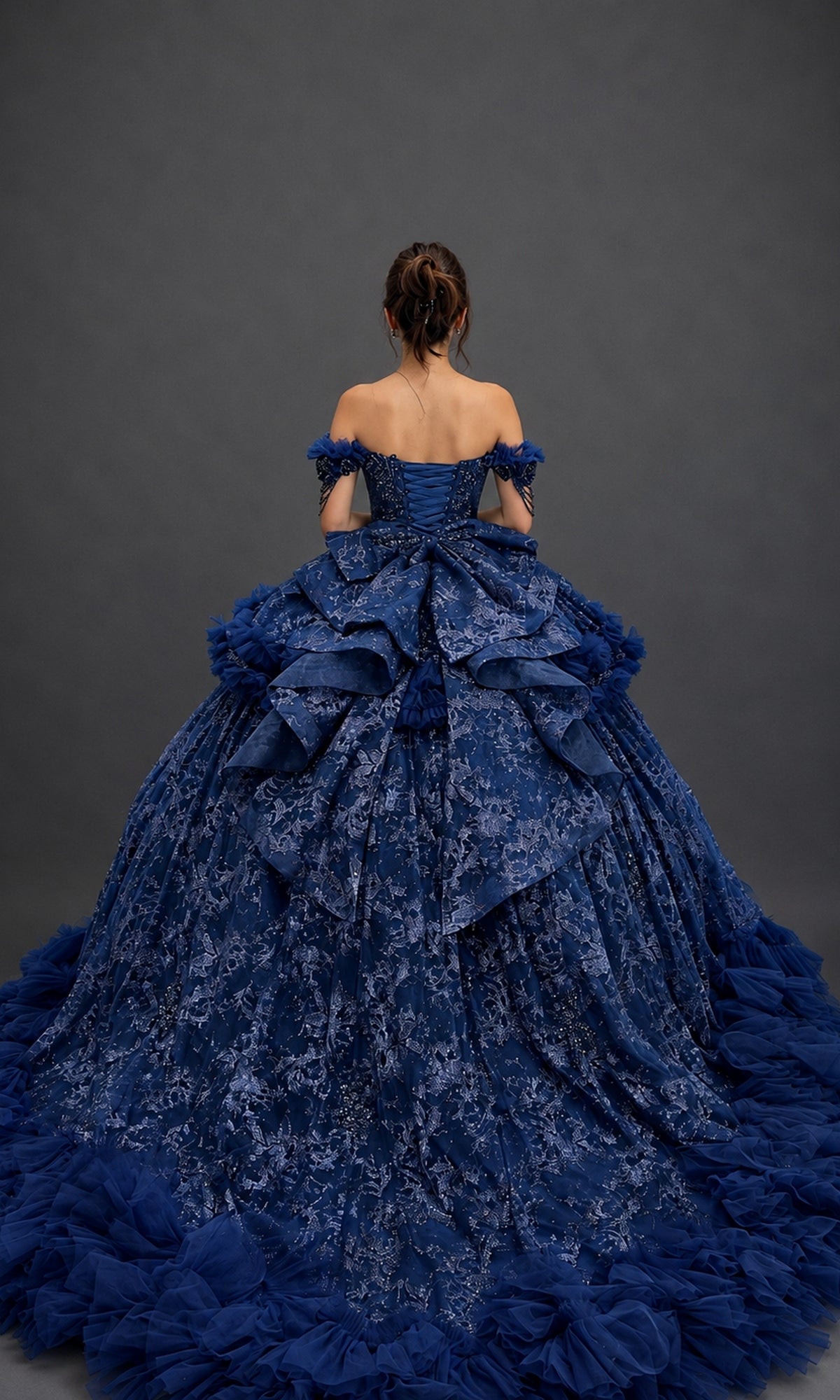 A woman stands facing away, wearing the Quinceanera Dress 5192 by Dancing Queen—a voluminous blue ball gown adorned with lace details, ruffles, and off-the-shoulder sleeves against a plain dark background. The shimmering beaded corset bodice adds an elegant touch to the timeless silhouette.